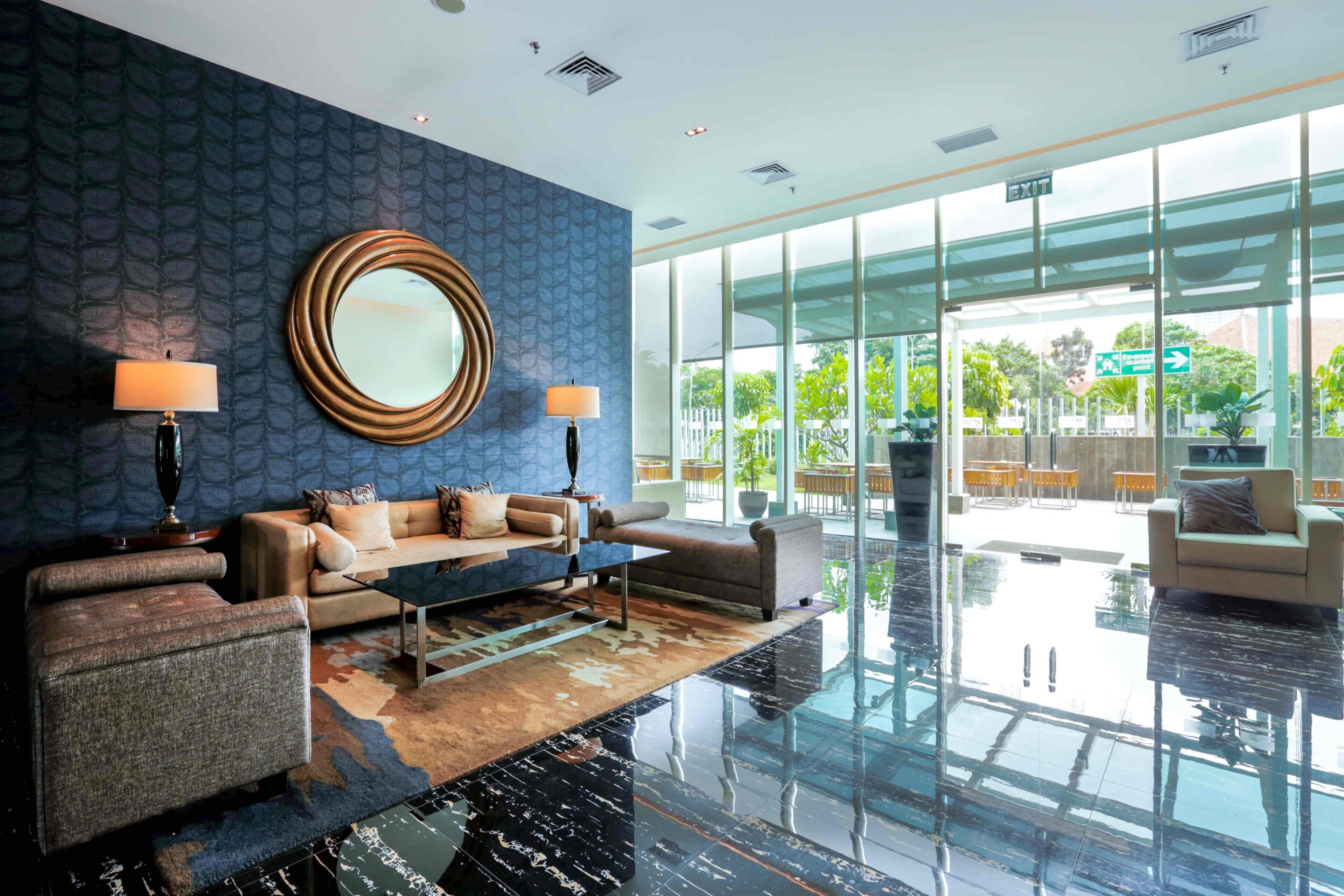 A modern lobby with a blue patterned wall, a round mirror, two sofas, and a glass entrance leading outside.