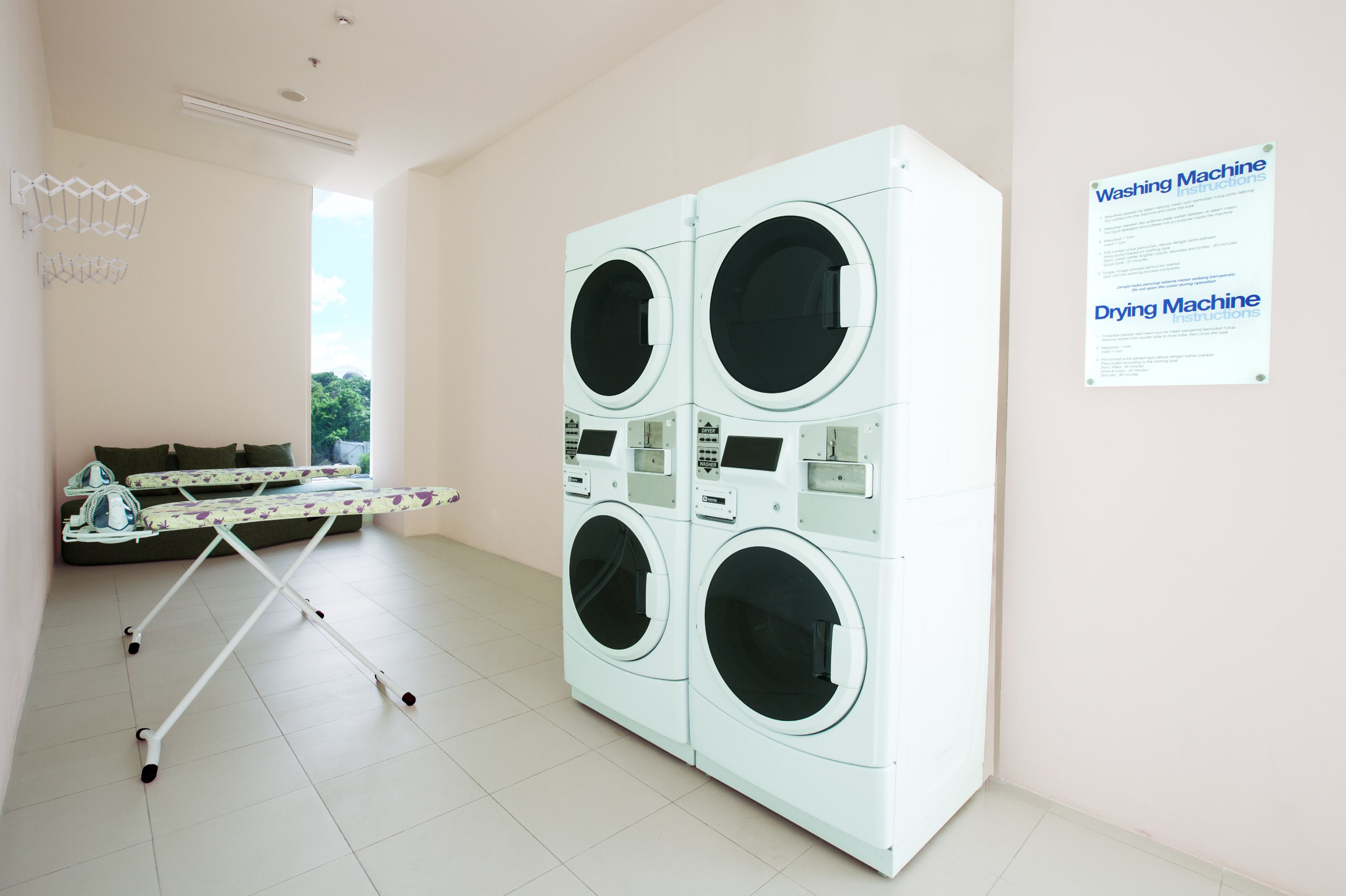 A laundry room with a large white washing and drying machine, a folding table with children's shoes, and a window providing natural light.