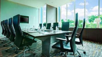 Un salón de conferencias con una mesa larga, varias sillas, un televisor de pantalla plana y ventanas amplias con vistas a zonas verdes.