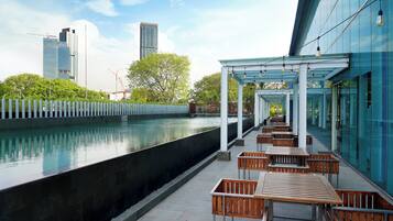 Un espace extérieur moderne, aménagé avec des tables et des chaises, situé près d’un point d’eau et offrant une vue sur un paysage urbain avec de hauts immeubles.
