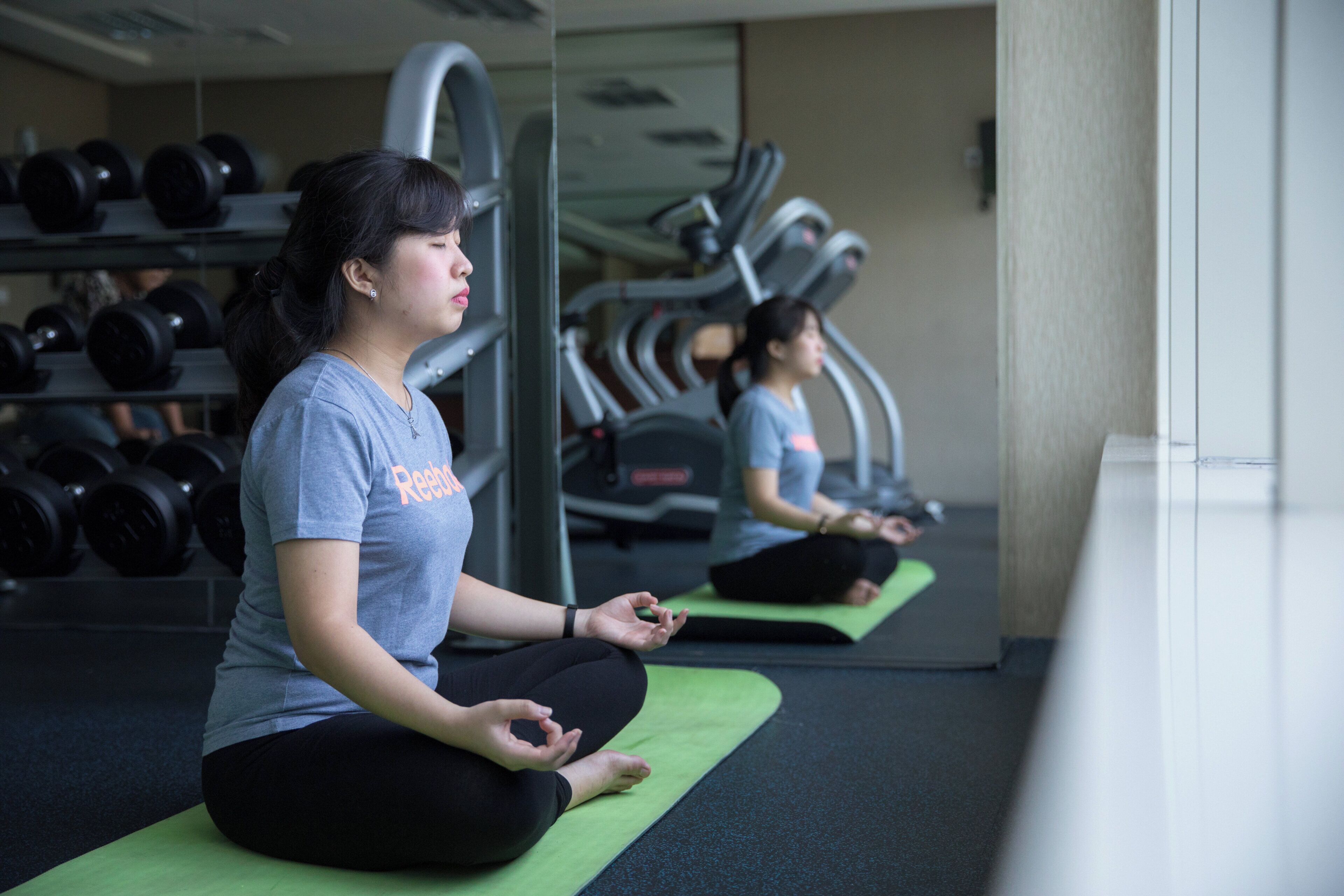 A person meditating on a green mat in a gym with exercise equipment in the background.