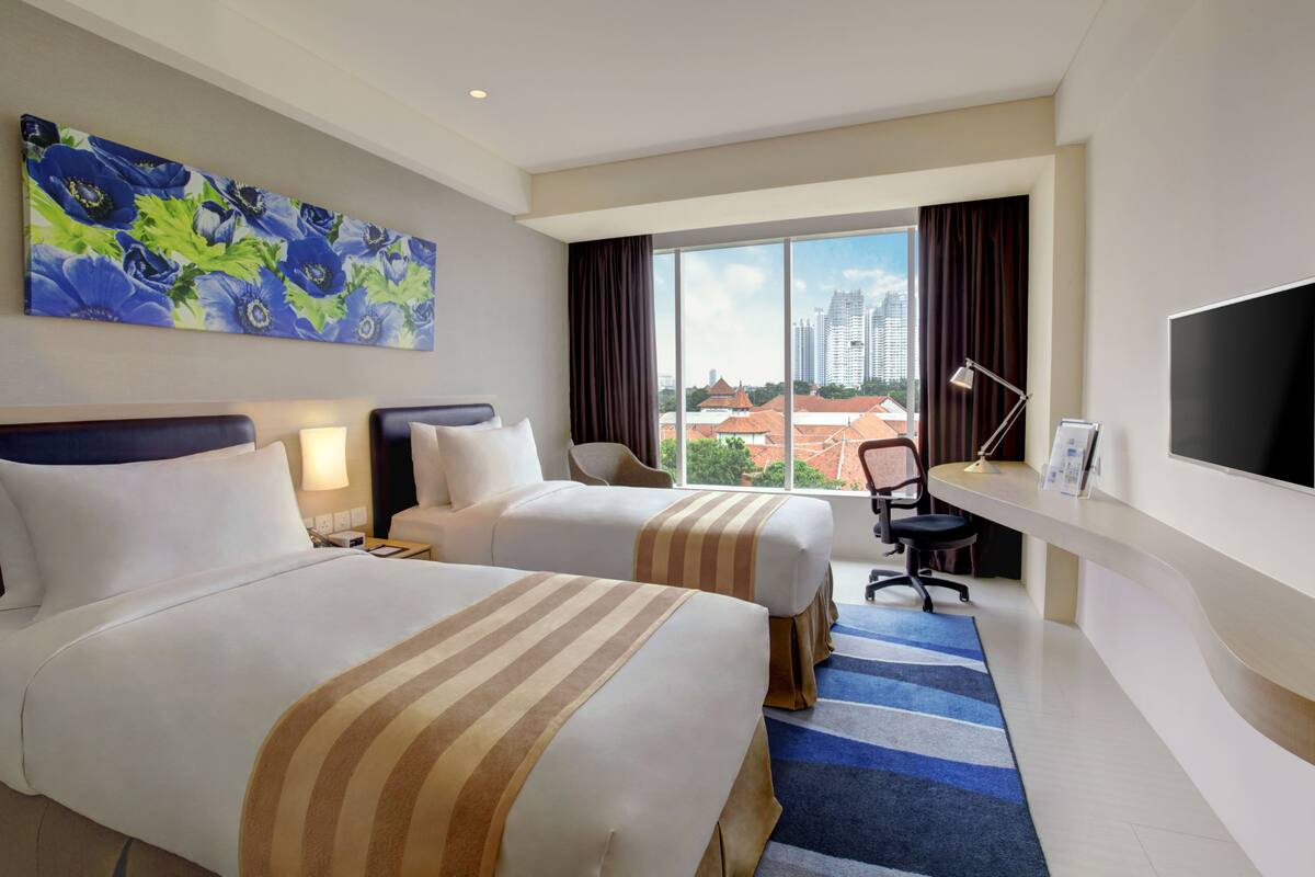 A hotel room with two beds, a large window with a city view, a flat-screen TV, and a desk with a lamp.