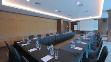 Un salón de conferencias con largas mesas, sillas, botellas de agua y una pantalla para proyector.
