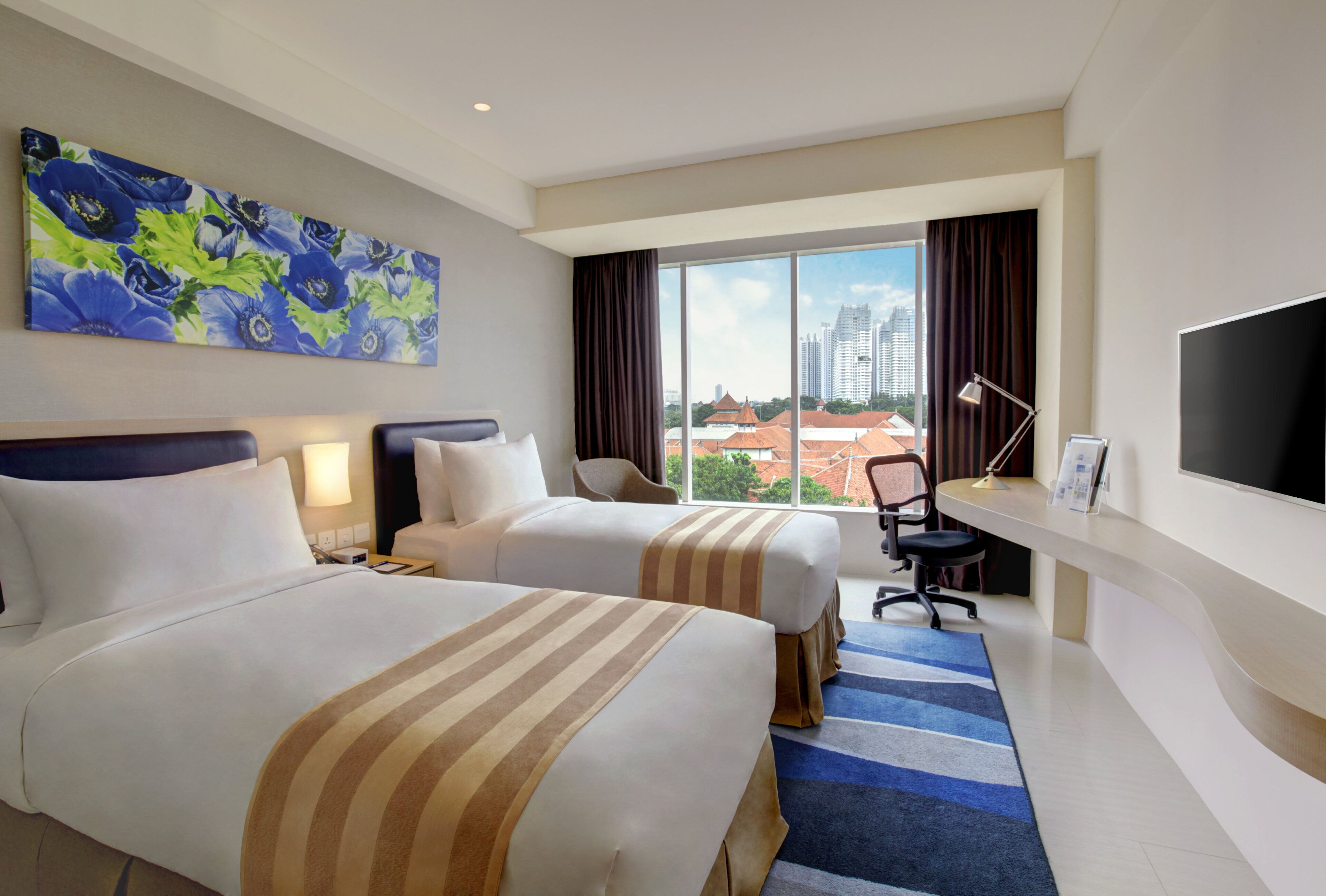 A hotel room with two beds, a large window with a city view, a flat-screen TV, and a desk with a chair.