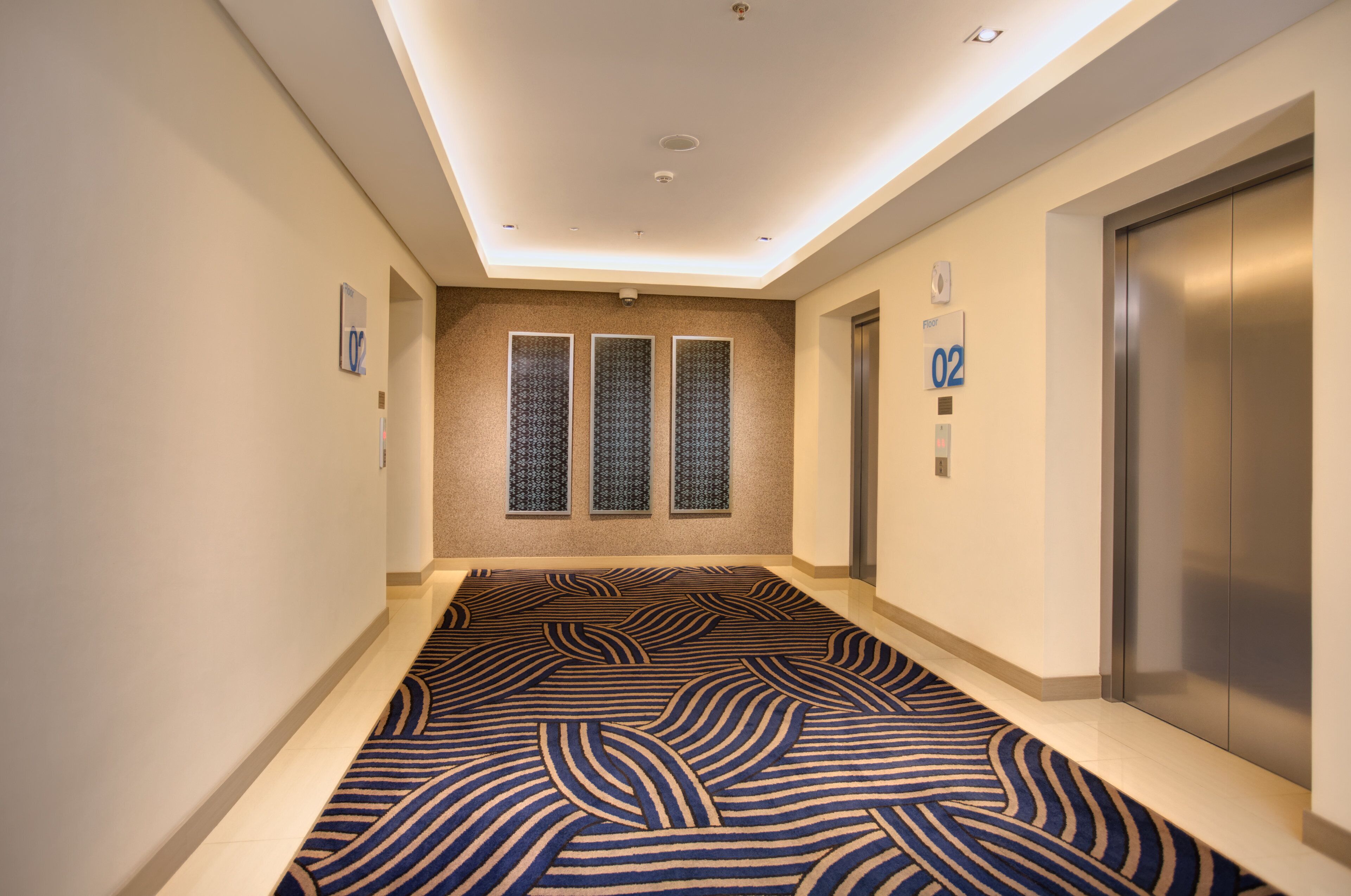 A hotel corridor with a patterned carpet, a wall-mounted elevator, and a ceiling with recessed lighting.