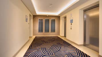 A hotel corridor with a patterned carpet, a wall-mounted elevator, and a ceiling with recessed lighting.