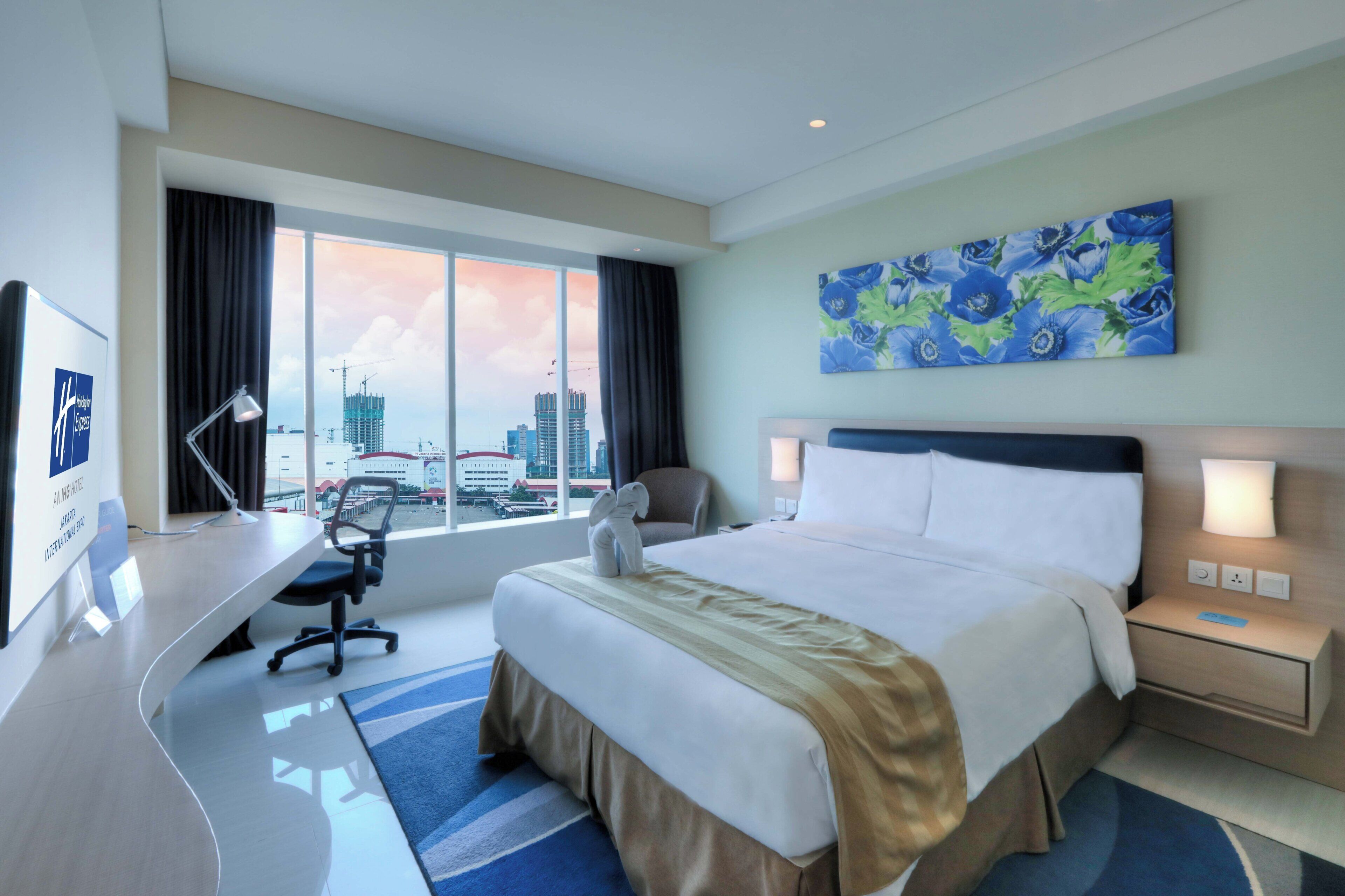 A hotel room with a bed, desk, chair, and a large window with a view of buildings.