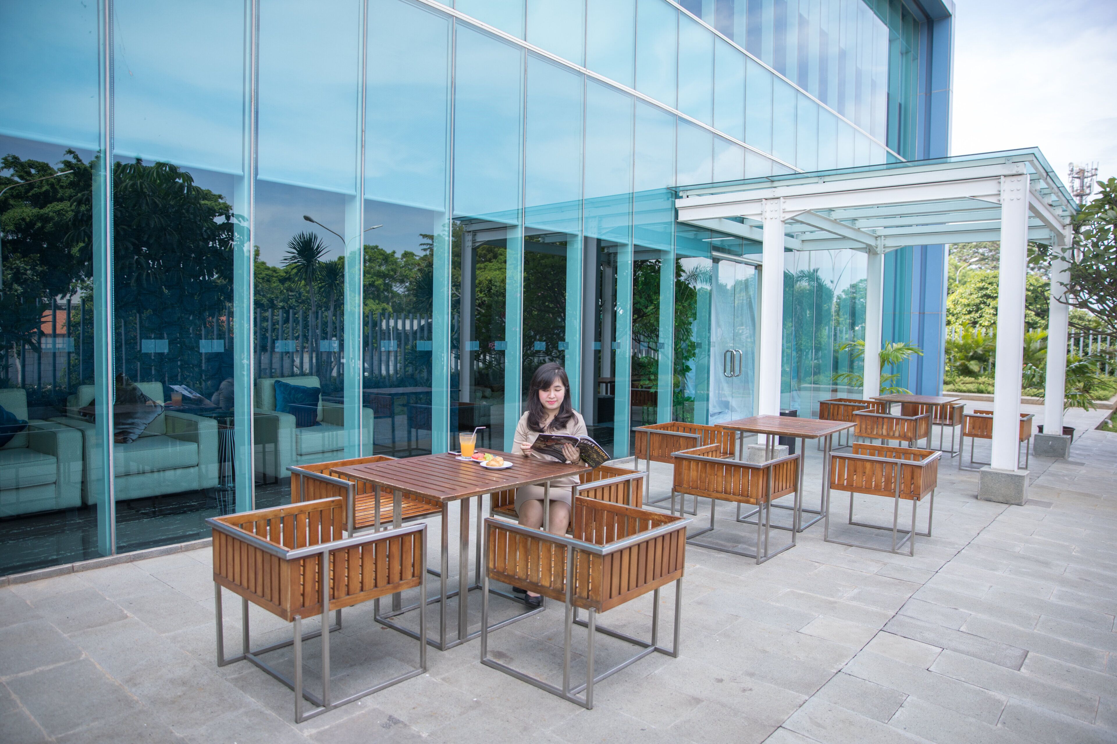 An outdoor seating area with tables and chairs, a glass building facade, and a person reading a magazine.