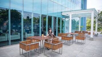 An outdoor seating area with tables and chairs, a glass building facade, and a person reading a magazine.