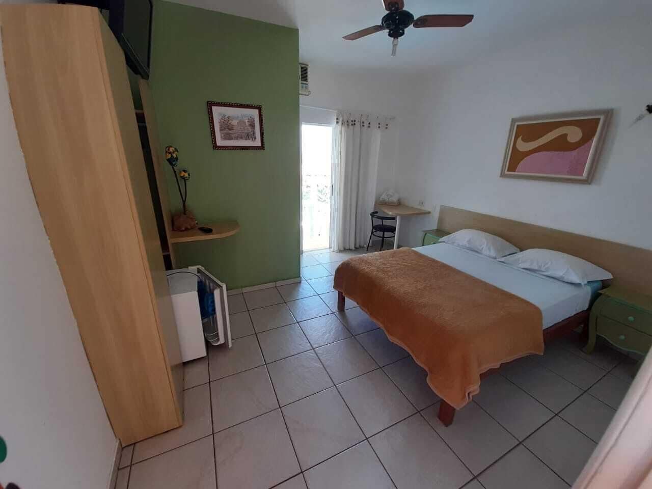 Un dormitorio con cama, mesita de noche, escritorio, silla, ventilador de techo y ventana con cortinas.