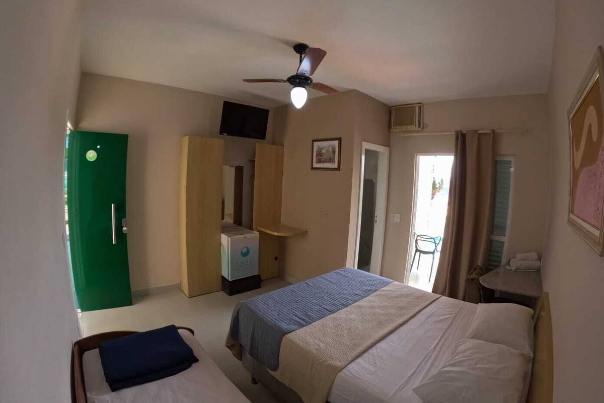 Quarto com duas camas, um ventilador de teto, uma porta verde, uma mesa pequena e uma cadeira.