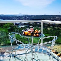 Un balcón con una mesa preparada para el desayuno, con vistas a un paisaje urbano y montañas al fondo.