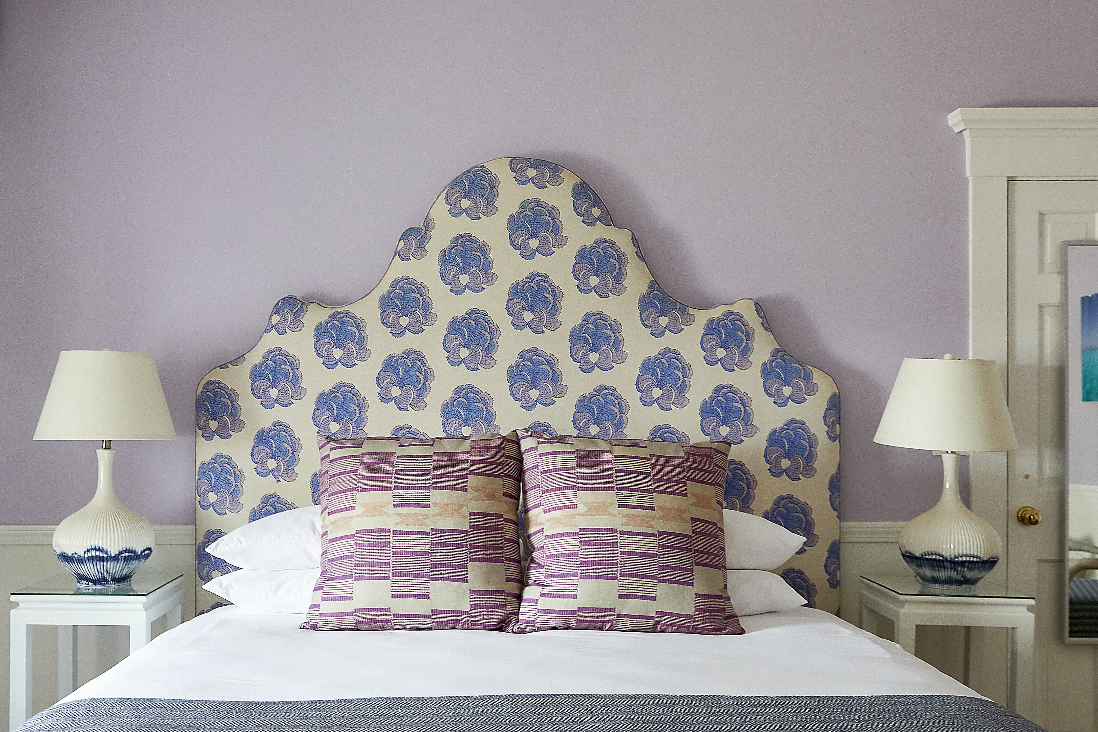 A neatly made bed with a patterned headboard, two bedside tables with lamps, and a wall with a floral wallpaper pattern.