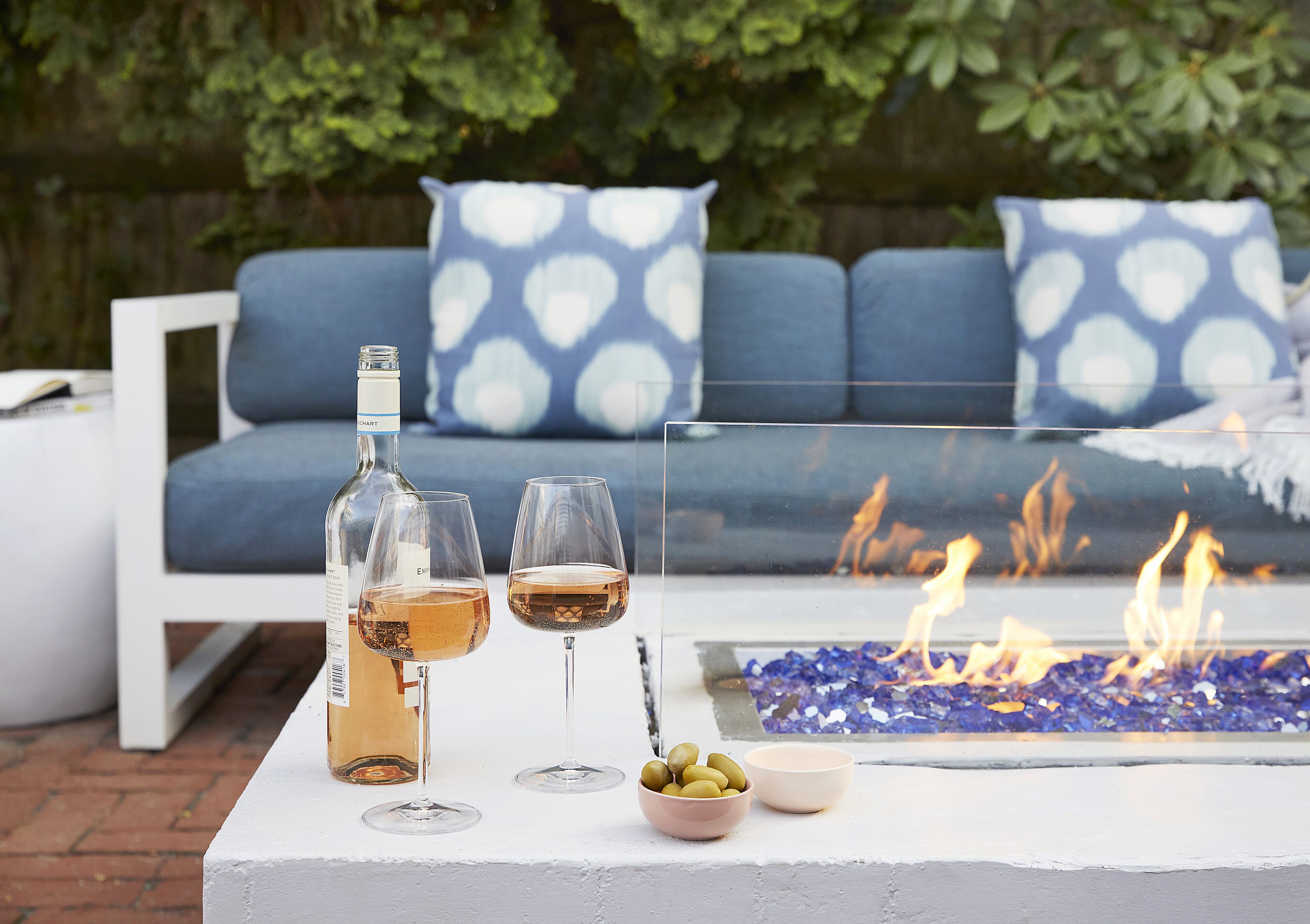 A fire pit with a glass table, two wine glasses, a bottle, and a bowl of olives.