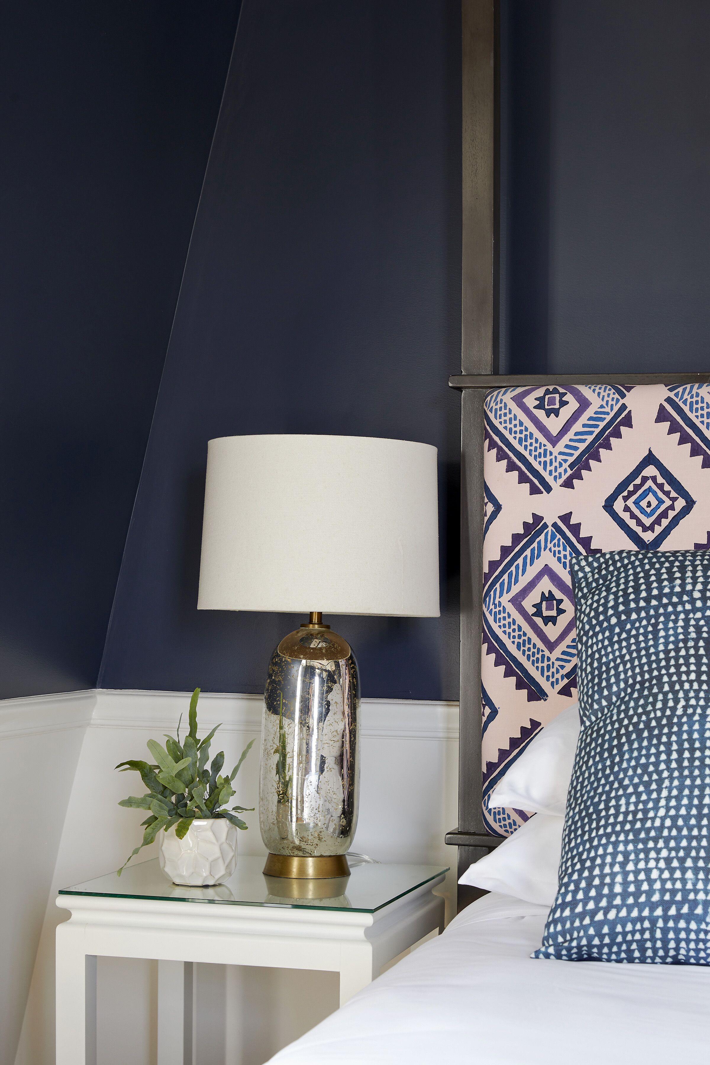 A neatly made bed with patterned pillows, a bedside lamp, and a small potted plant on a glass-topped nightstand.