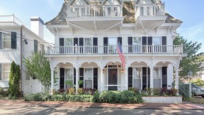 A two-story white colonial-style house with a balcony, a front porch, and an American flag. - The Christopher, The Edgartown Collection (Edgartown)