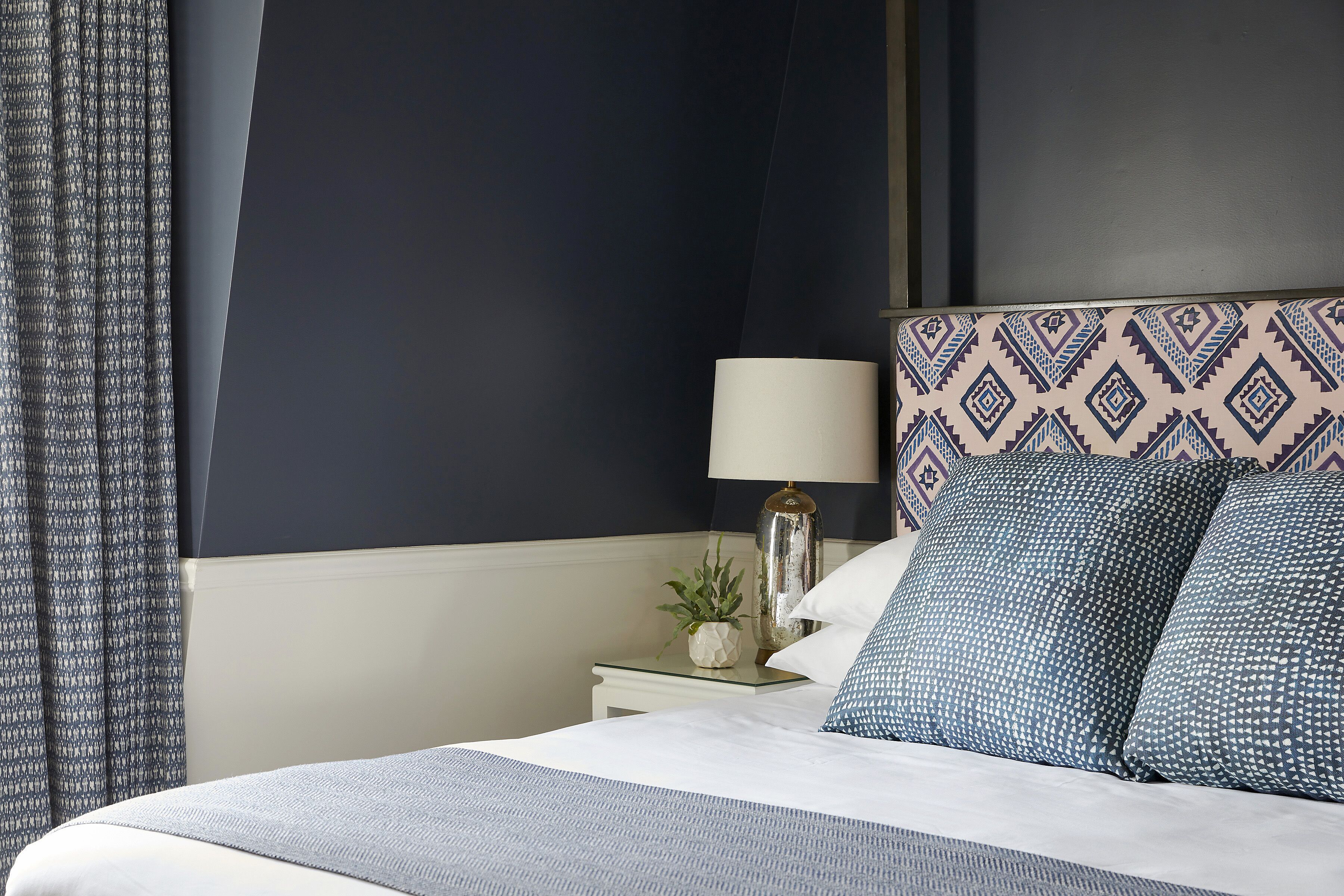A neatly made bed with a patterned headboard, a bedside lamp, and a small plant on a nightstand.