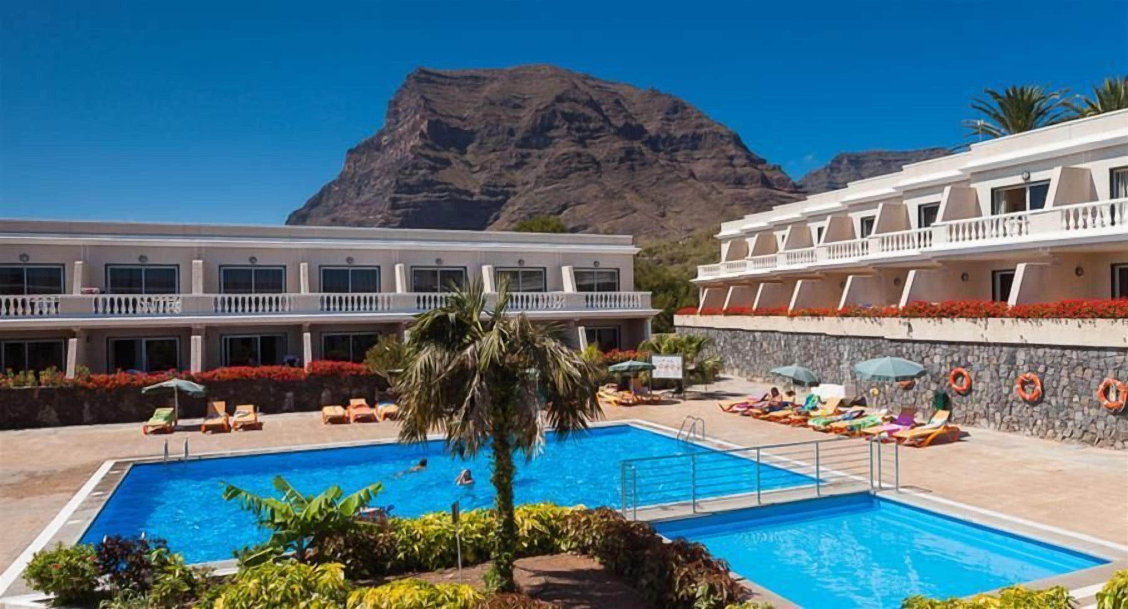 Hotel amb piscina, gandules i muntanyes al fons.