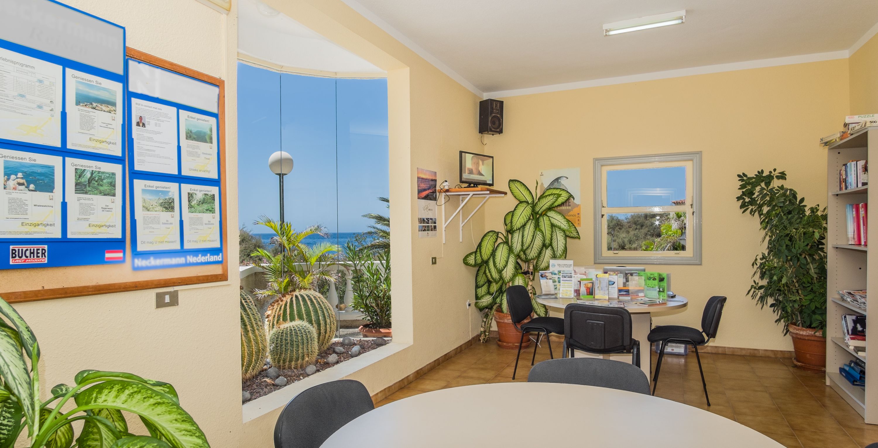 A room with a large window, a bulletin board with papers, a table with chairs, and a bookshelf.
