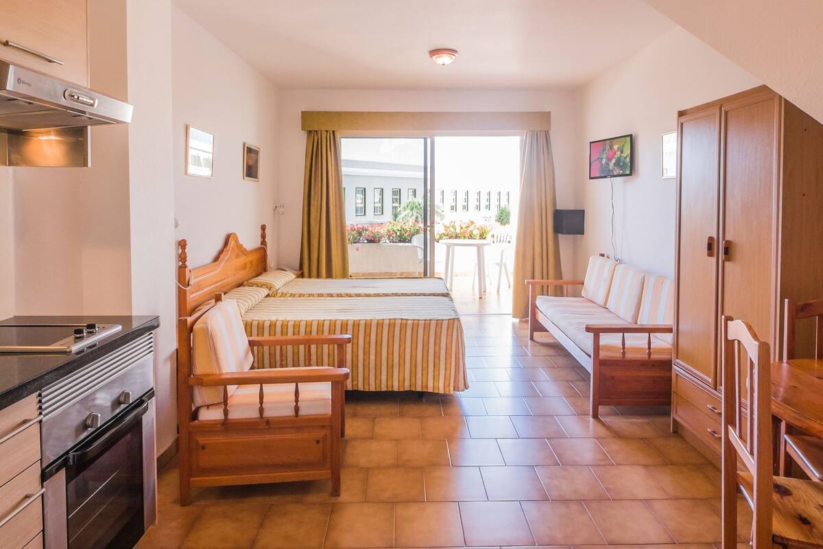 Quarto de hotel com cozinha compacta, cama com roupa de cama listrada, uma cadeira de madeira, um sofá e uma televisão na parede.