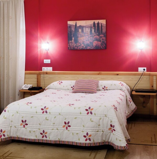 A hotel room with a red wall, a painting, a floral bedspread, a striped pillow, and bedside lamps.