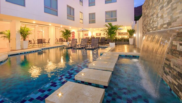 A hotel pool area with a tiled pool, lounge chairs, and a waterfall feature.