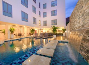 Ett poolområde vid hotellet med en kakelbeklädd pool, solstolar och en dekorativ vattenfallsanläggning.