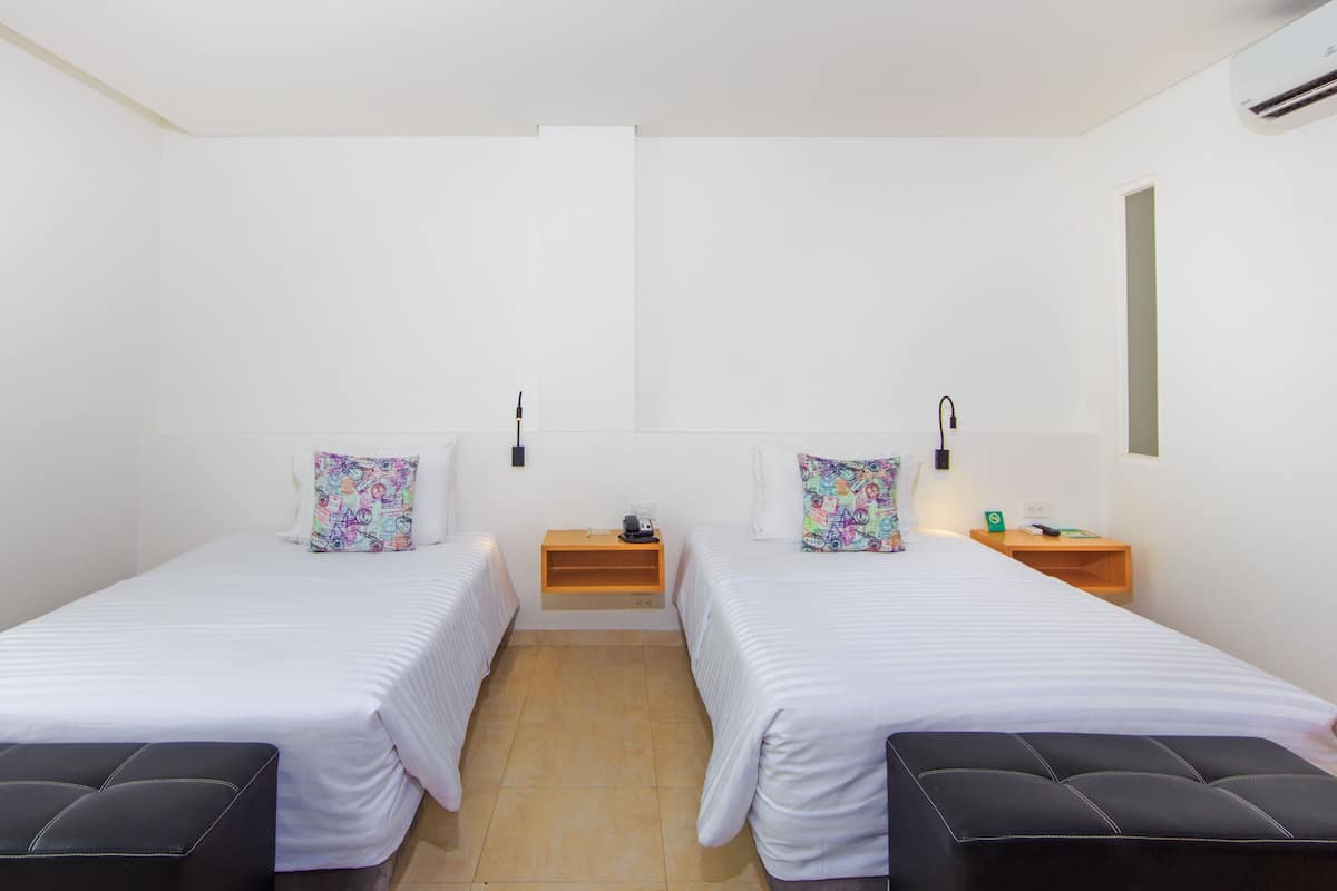 Habitación doble con dos camas, cada una con un cojín de colores, una mesita de noche con lámpara y un pequeño estante.
