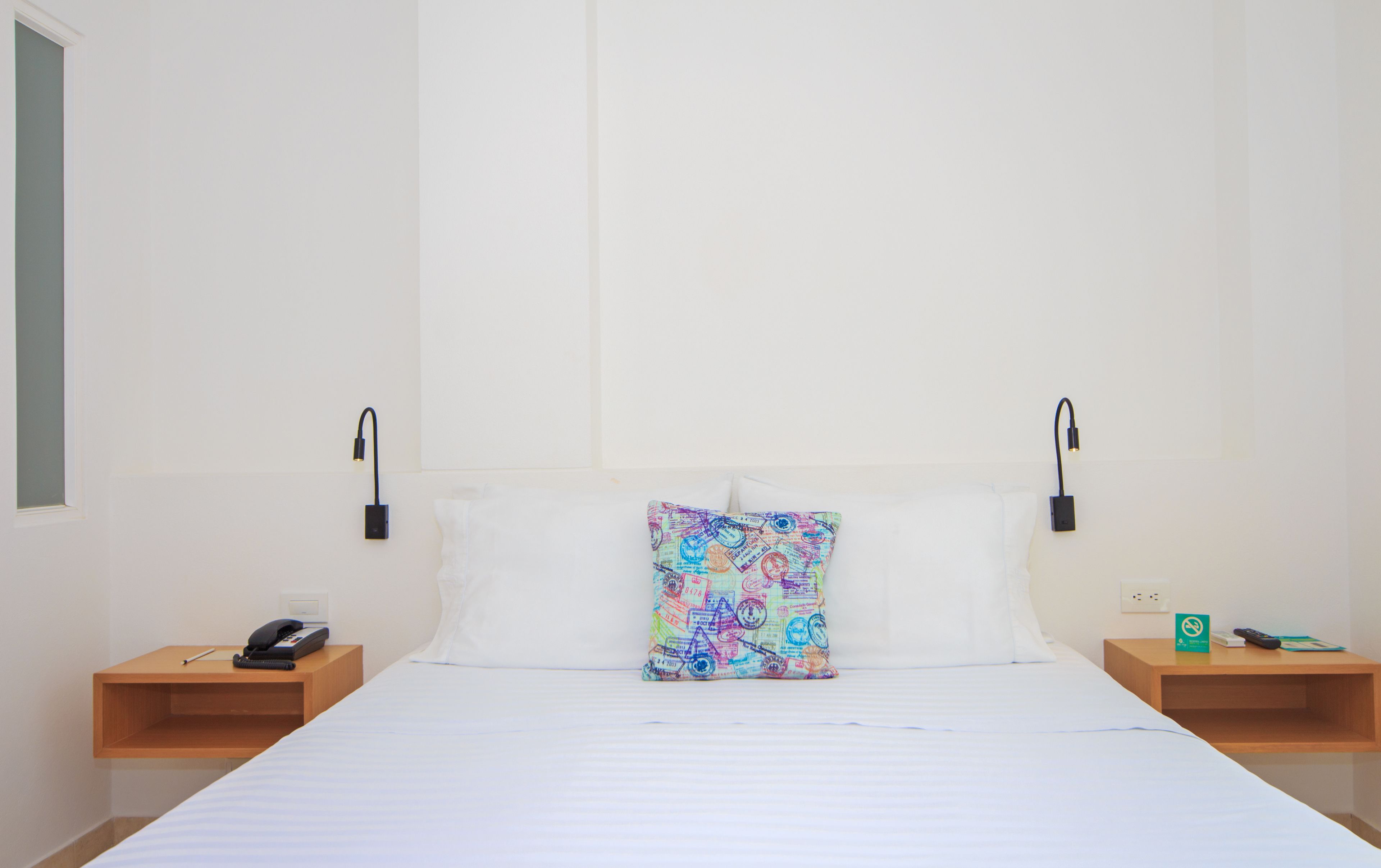 A hotel room with a bed, two bedside tables, a nightstand, a pillow, and a wall-mounted lamp.