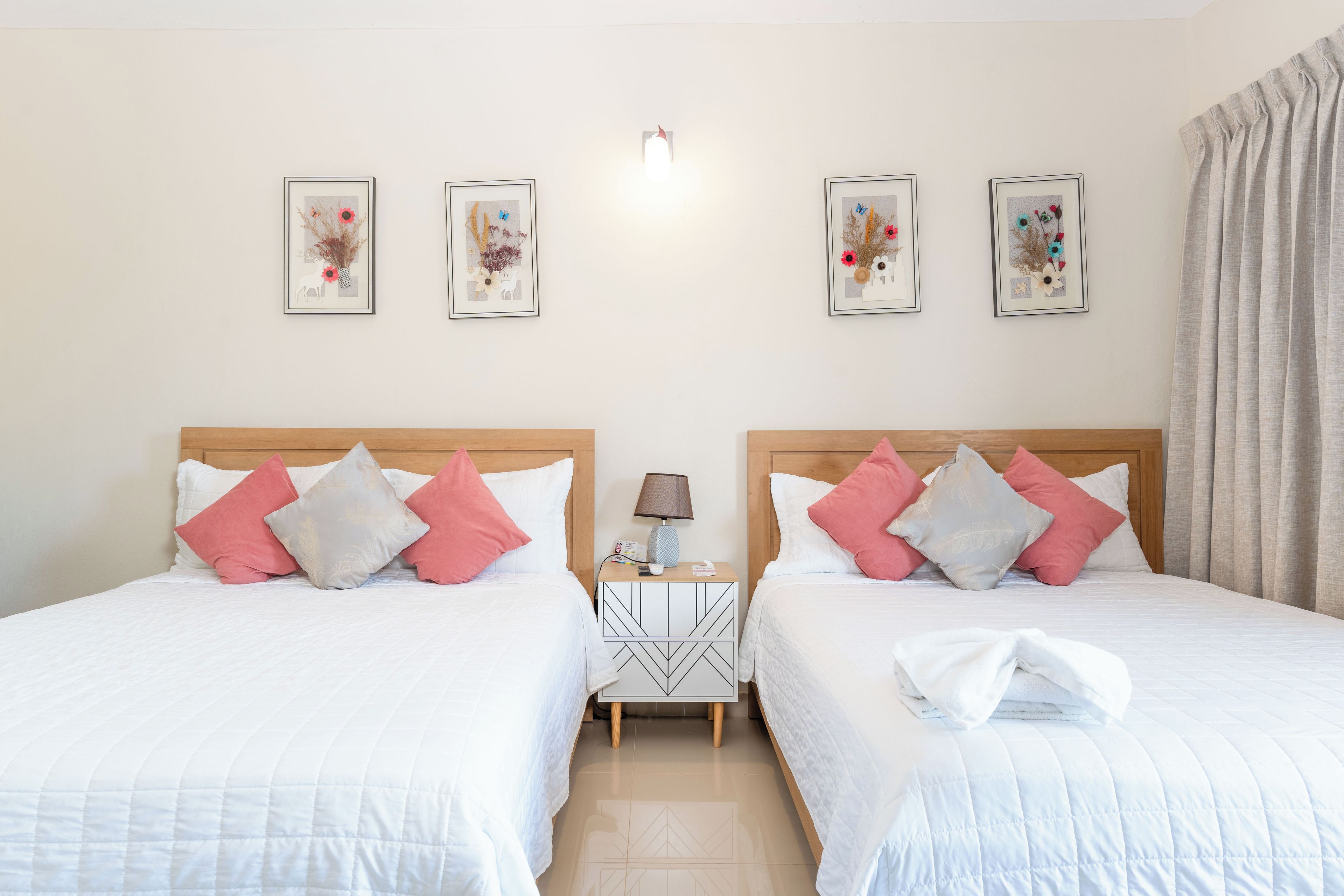 A hotel room with two beds, each with white bedding and pink and grey pillows, a bedside table with a lamp, and three framed floral artworks on the wall.