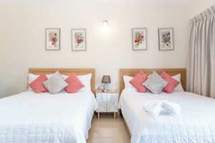 Habitación de hotel con dos camas, cada una con ropa blanca y almohadas rosa y gris, una mesita de noche con lámpara, y tres cuadros con motivos florales en la pared.