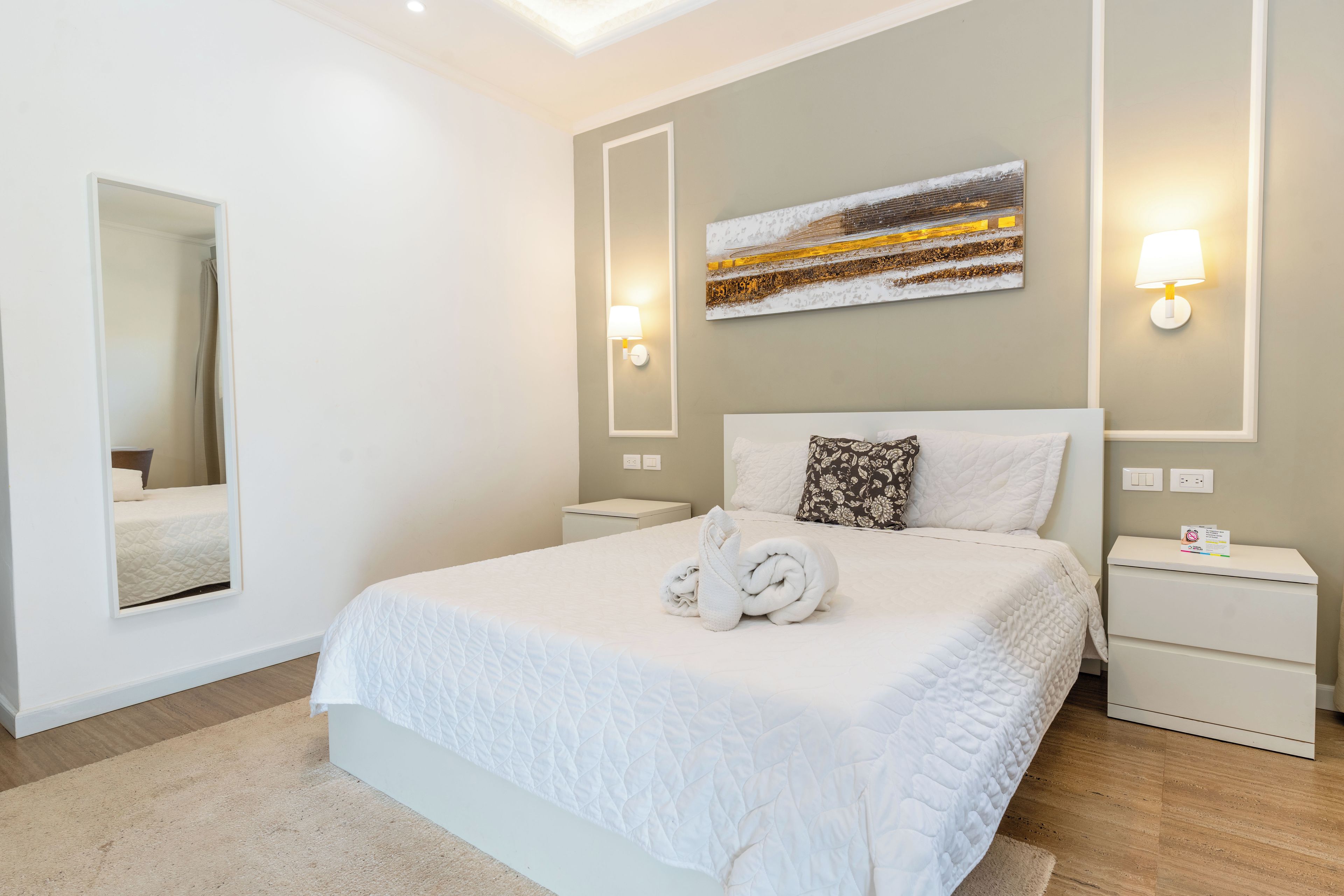 A hotel room with a large bed, two bedside tables, a wall-mounted lamp, a mirror, and a painting.