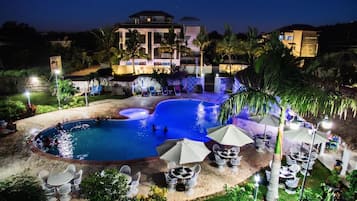 Vista noturna de um resort com piscina, espreguiçadeiras e um prédio com uma placa.