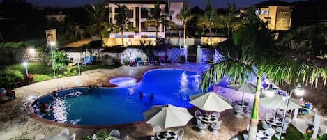 A nighttime view of a resort with a swimming pool, lounge chairs, and a building with a sign.