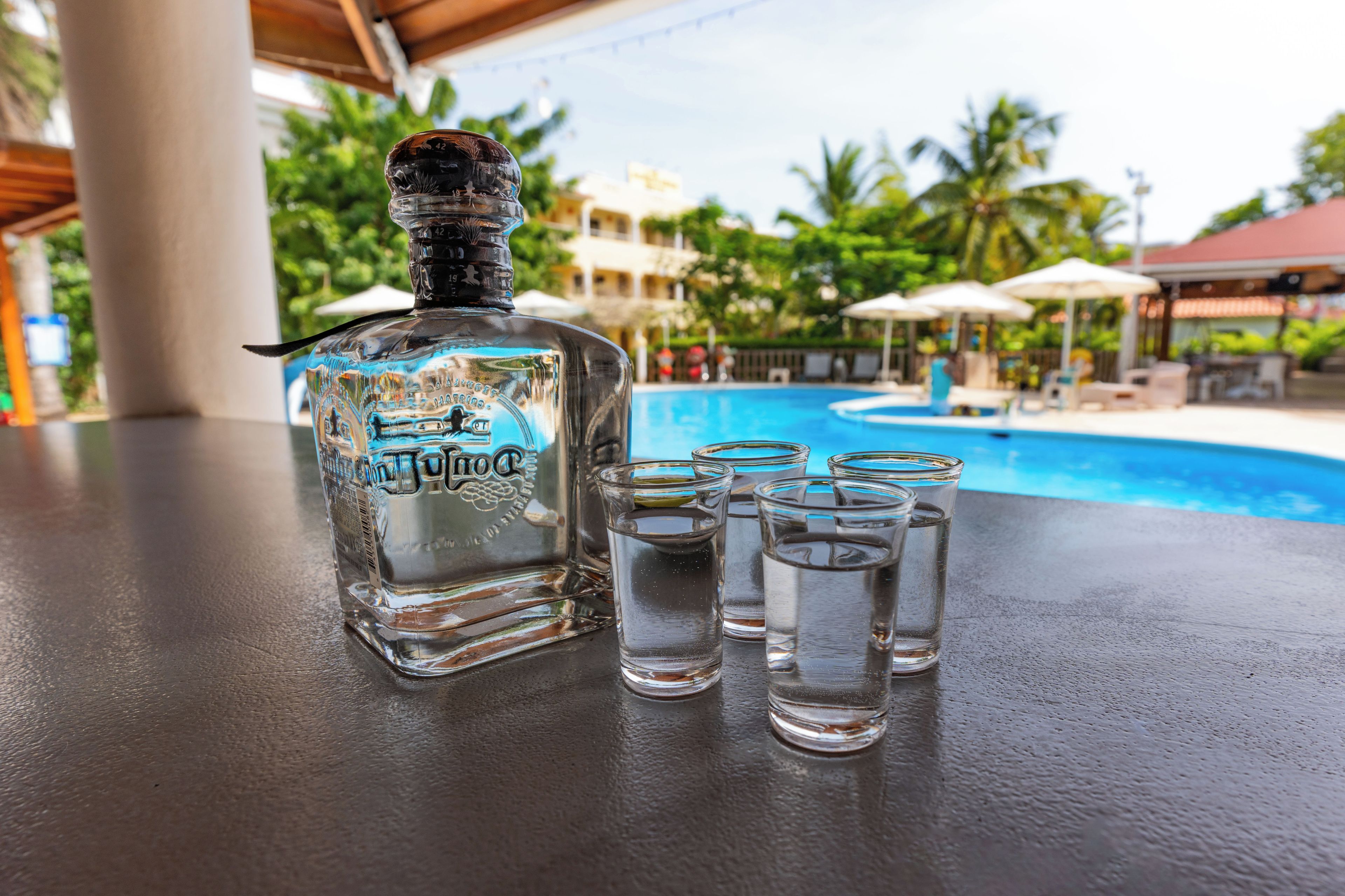A bottle of liquor and shot glasses on a table with a pool in the background.
