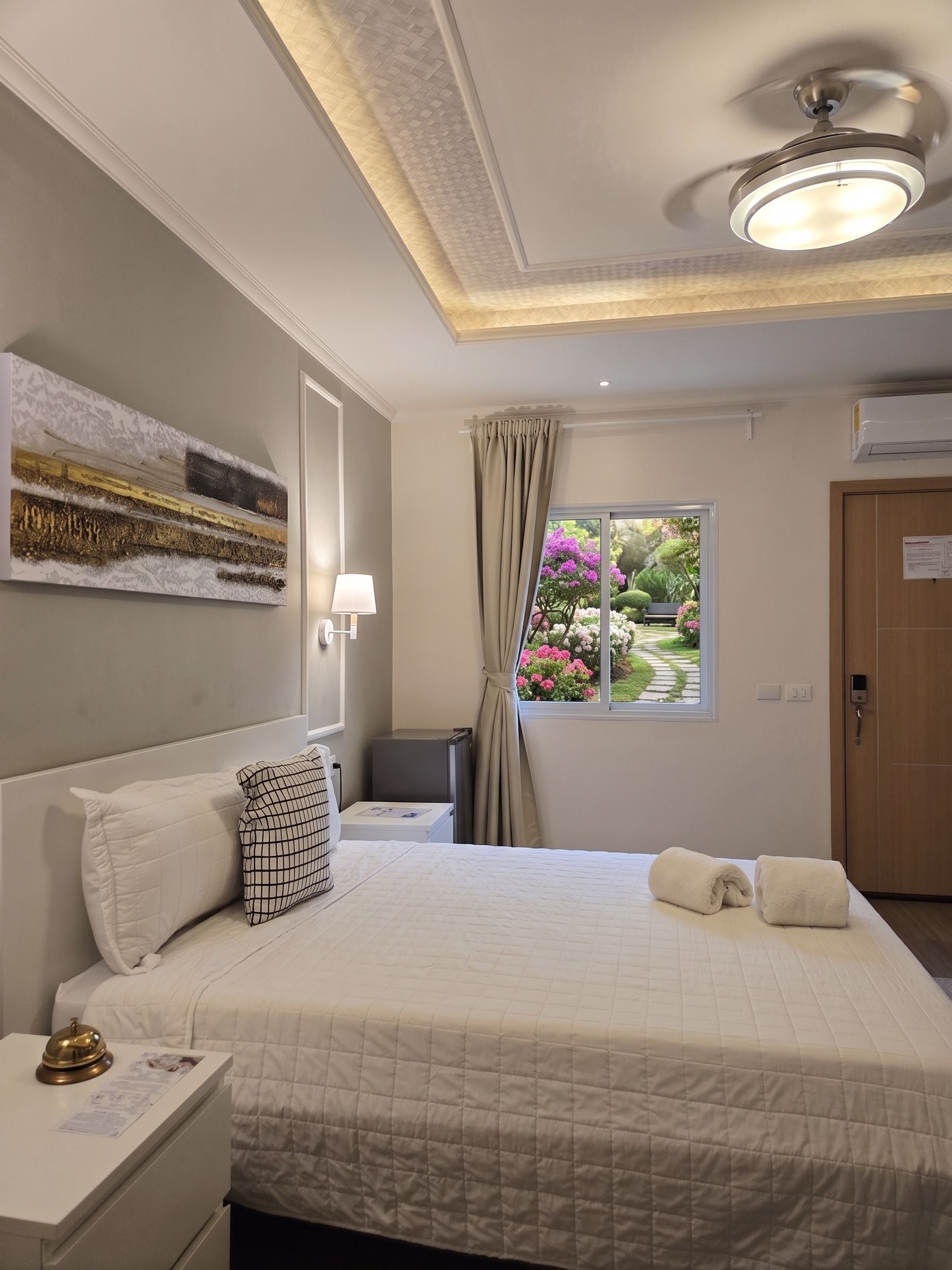 A hotel room with a bed, a bedside table, a lamp, a window with a view of a garden, and a door.