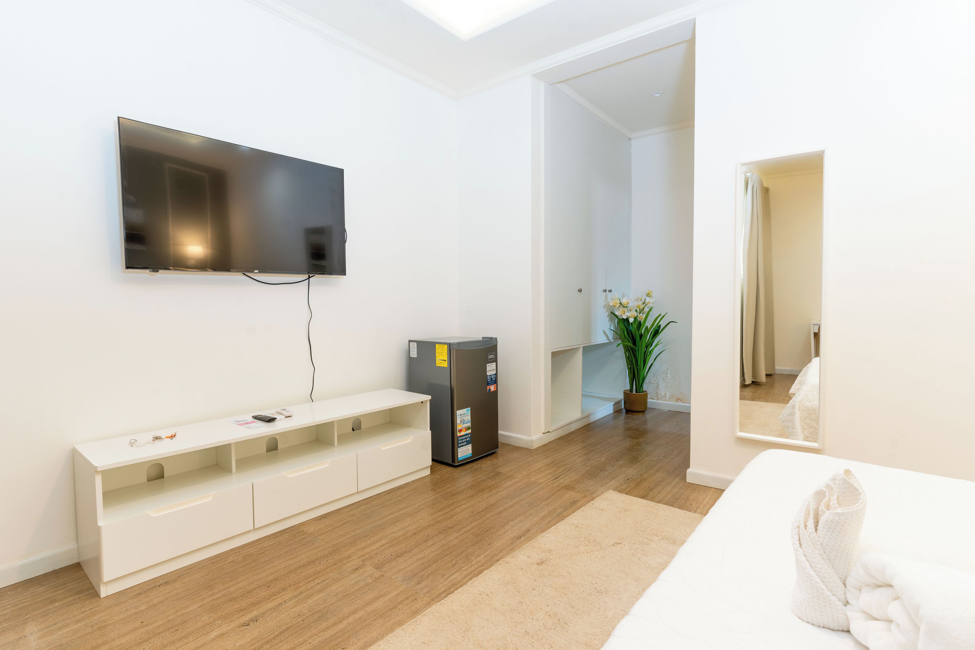 Et moderne hotelværelse med et vægmonteret fladskærms-tv, en indbygget bænk med opbevaring, et lille køleskab og en potteplante.