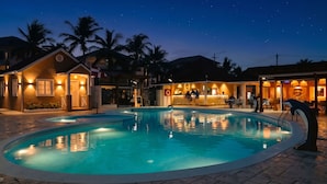 A nighttime pool area with a bar and lounge seating, surrounded by palm trees and buildings. - Casual Eden Bayahibe by Casual Hoteles (Dominicus)