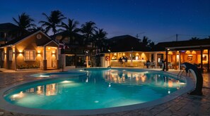 A nighttime pool area with a bar and lounge seating, surrounded by palm trees and buildings. - Casual Eden Bayahibe by Casual Hoteles (Dominicus)