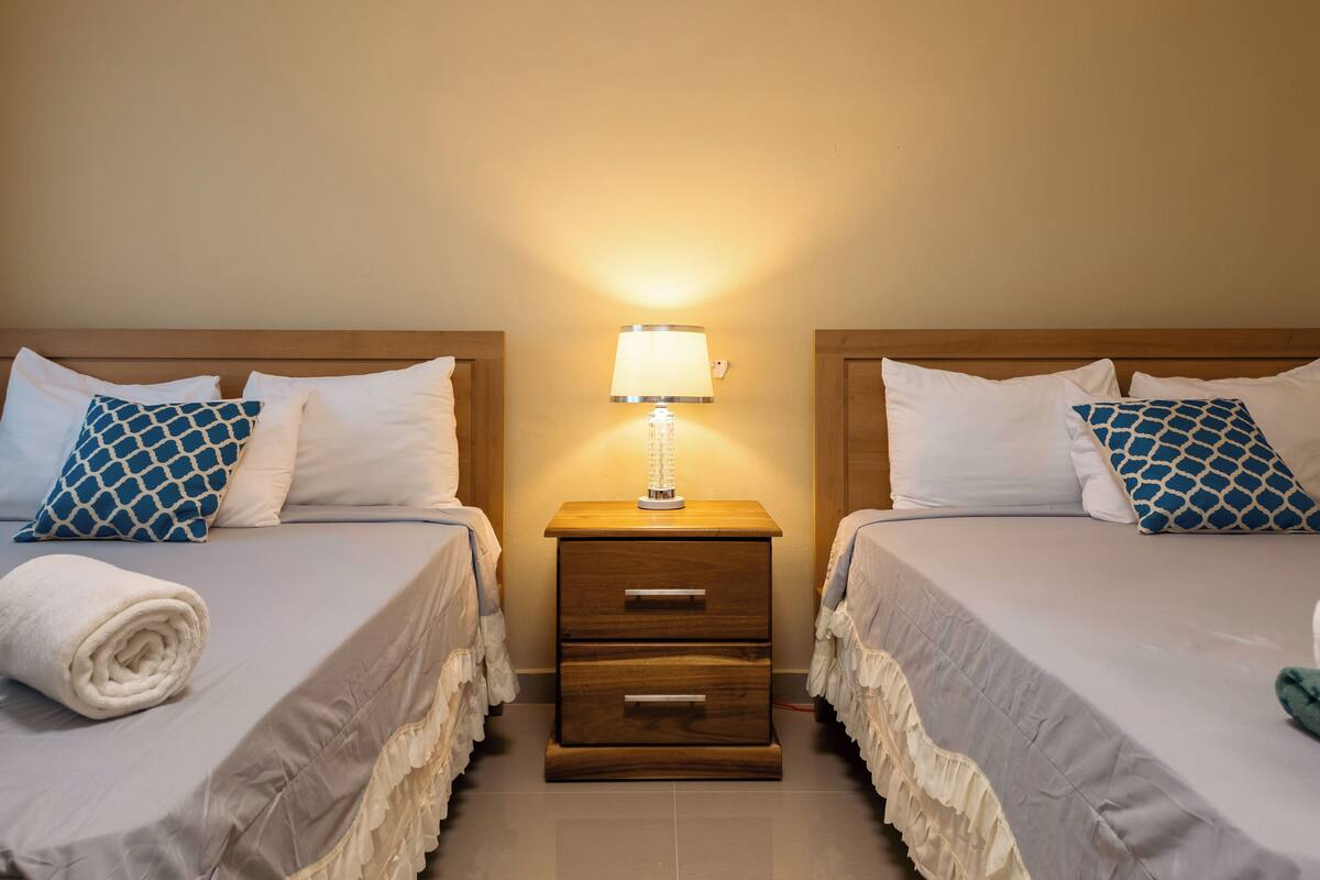 A hotel room with two beds, each with a white and blue patterned pillow, a wooden nightstand with a lamp, and a rolled towel on the left bed.