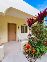 Entrada de un hotel con puerta de madera, una silla blanca y una maceta con plantas y flores tropicales.