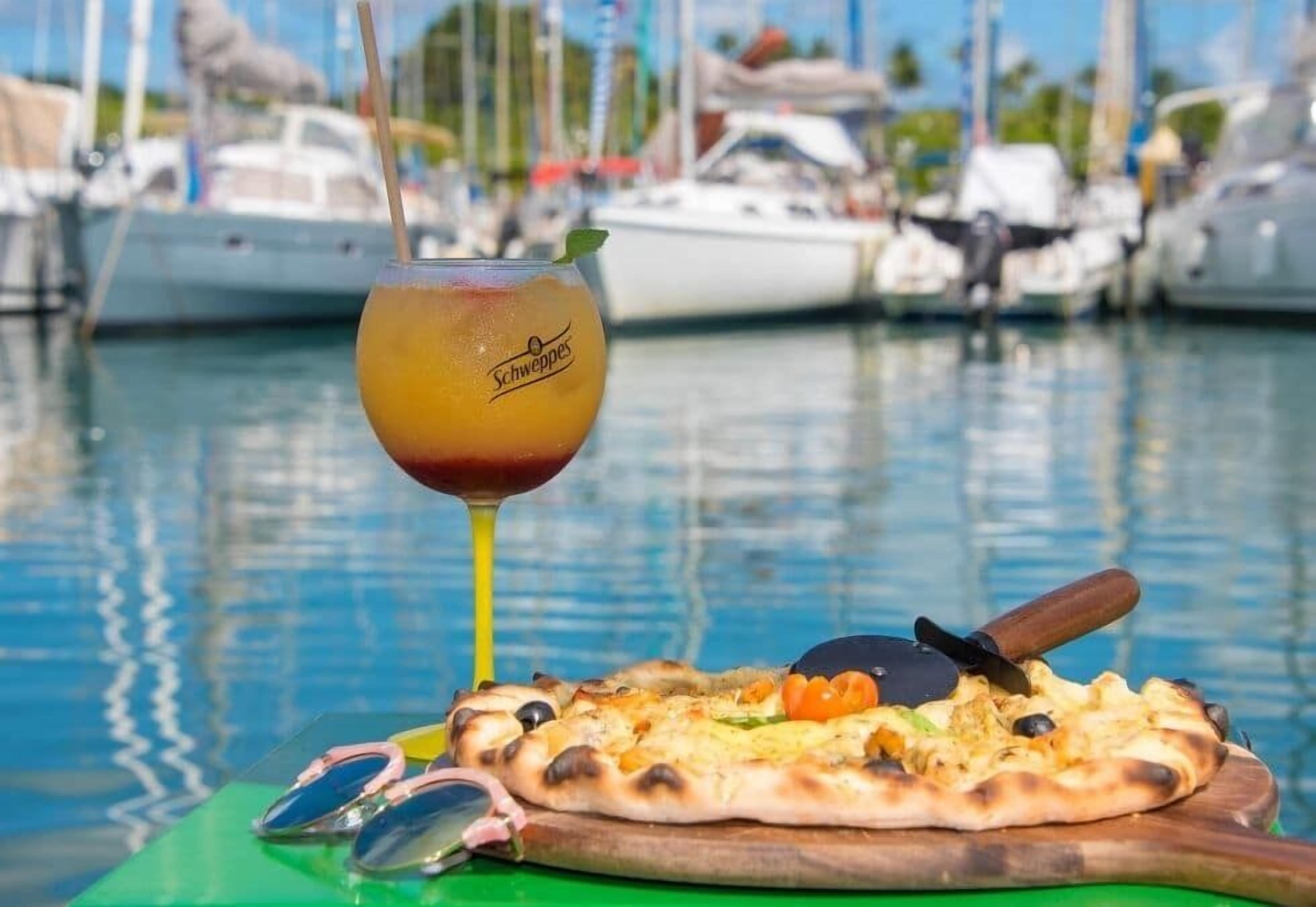 A pizza with a pizza cutter on a green cutting board, a glass of drink with a straw, and a pair of sunglasses on a green surface.