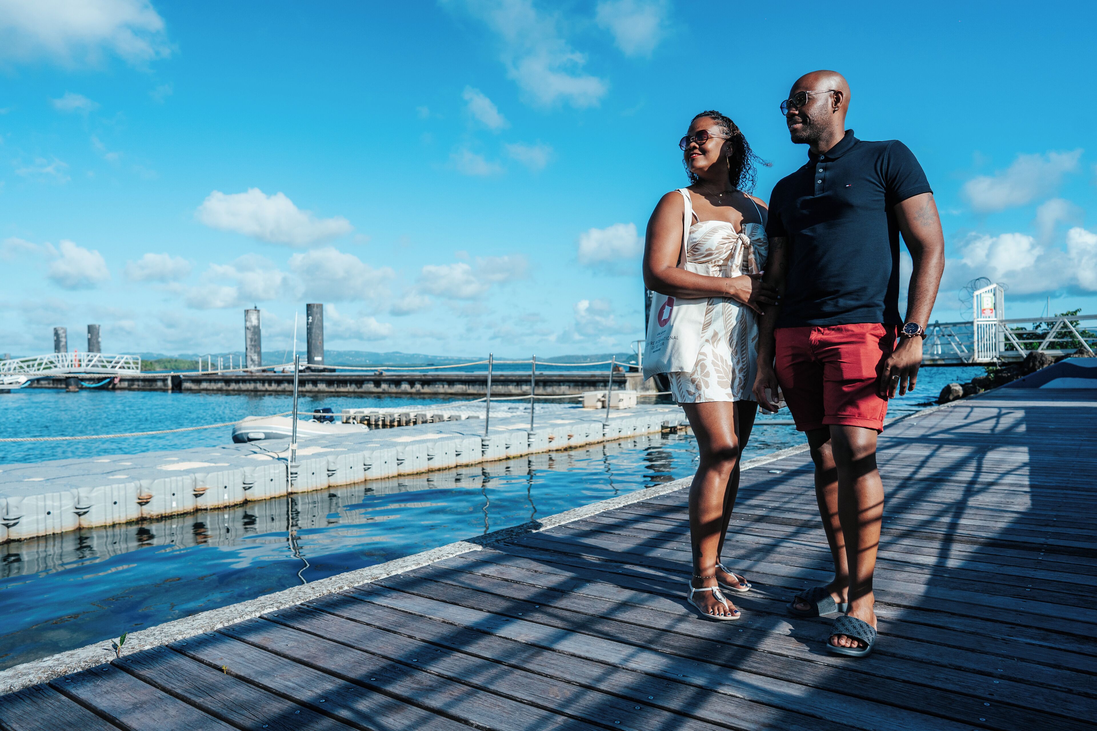 Un couple se tient sur une jetée en bois, avec une vue sur une marina et un pont en arrière-plan.