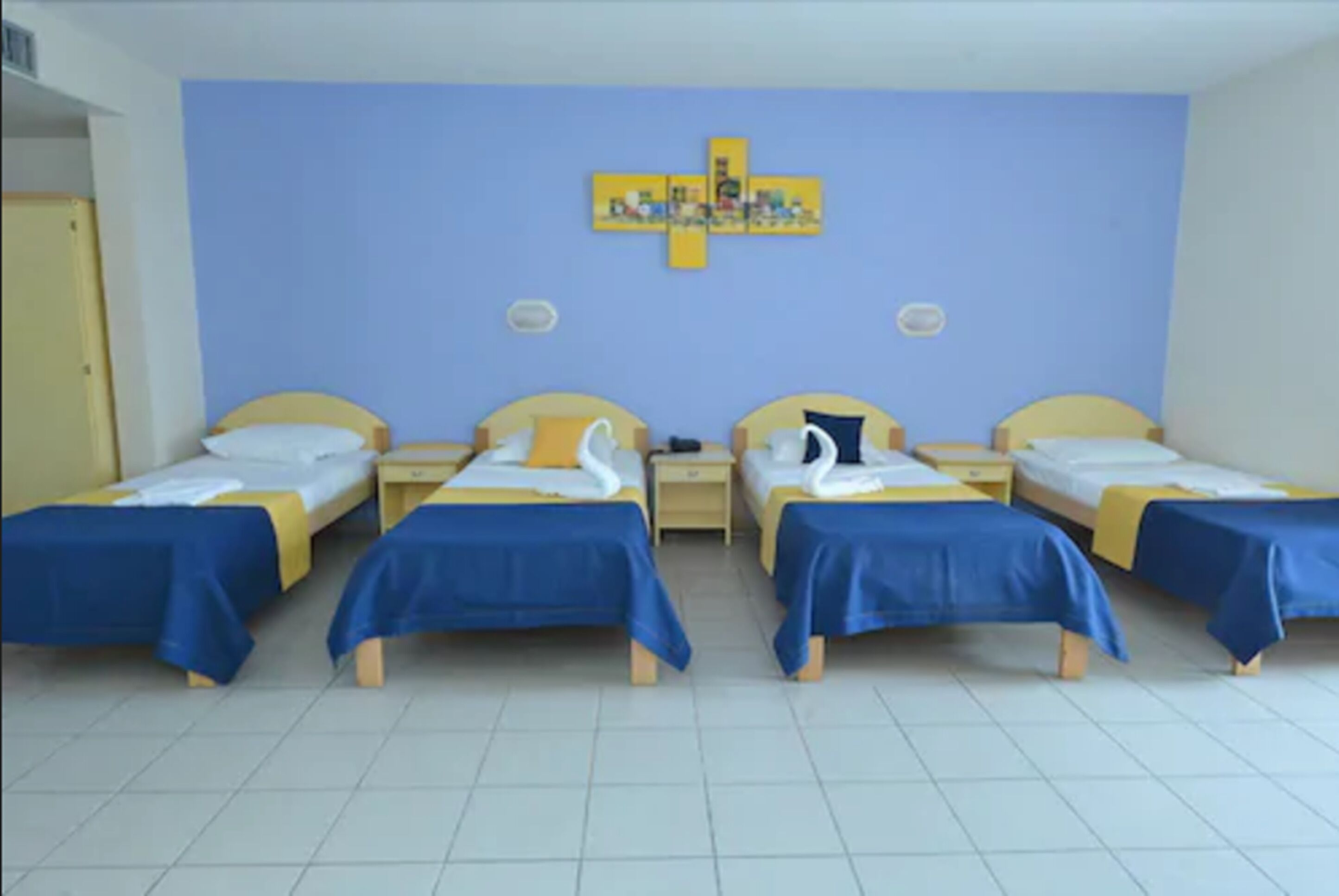 Une chambre d’hôtel avec quatre lits simples, chacun recouvert d’une couverture bleue et agrémenté d’oreillers blancs. Un mur bleu de la chambre est orné d’une œuvre d’art jaune, placée au-dessus des lits.