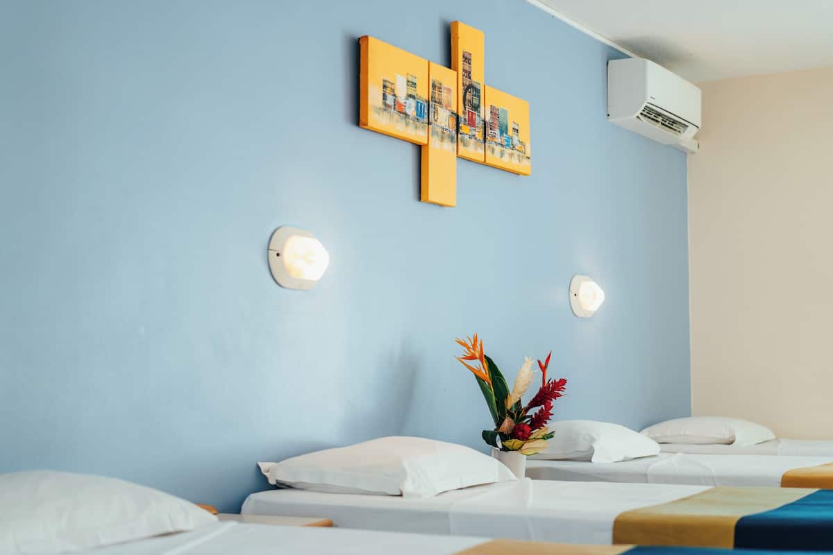 Una habitación de hotel con una pared azul, una obra de arte con forma de cruz, dos mesitas de noche con lámparas y un arreglo floral.
