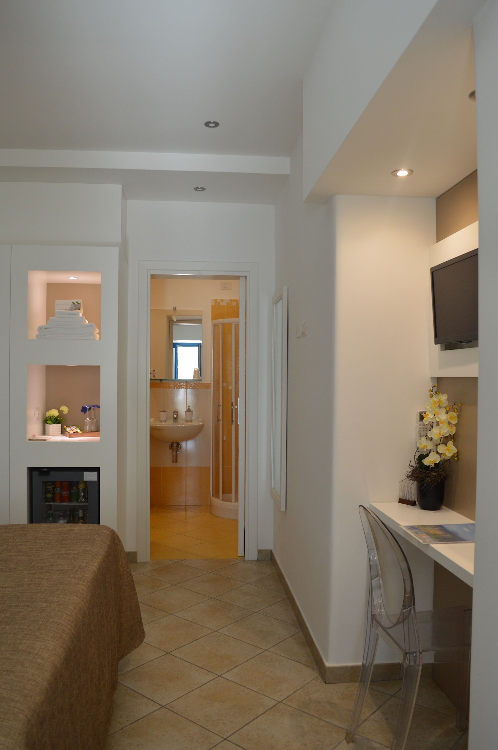 A hotel room with a bed, a desk with a chair, a TV, a bathroom with a sink, and a small shelf with flowers.