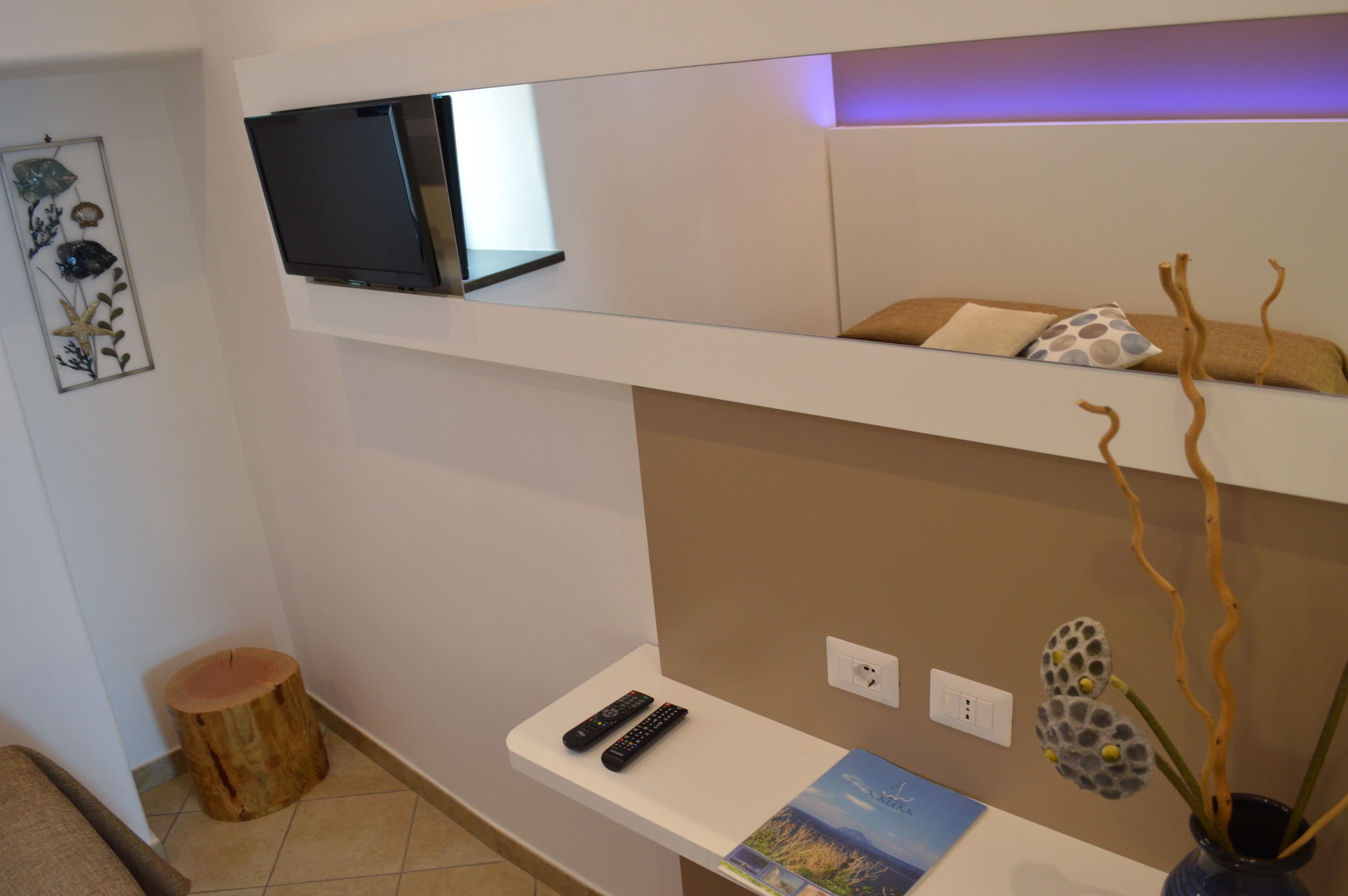 A modern room with a flat-screen TV on a white shelf, a small table with a remote control, a decorative plant, and a wall-mounted outlet.