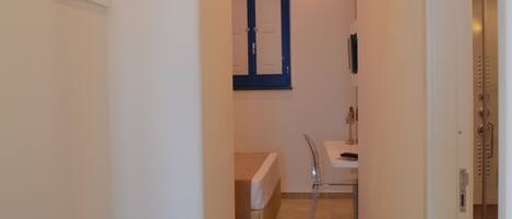 Un couloir étroit, avec un banc sur le côté gauche, qui mène à une salle de bain équipée d’un miroir et d’un lavabo.