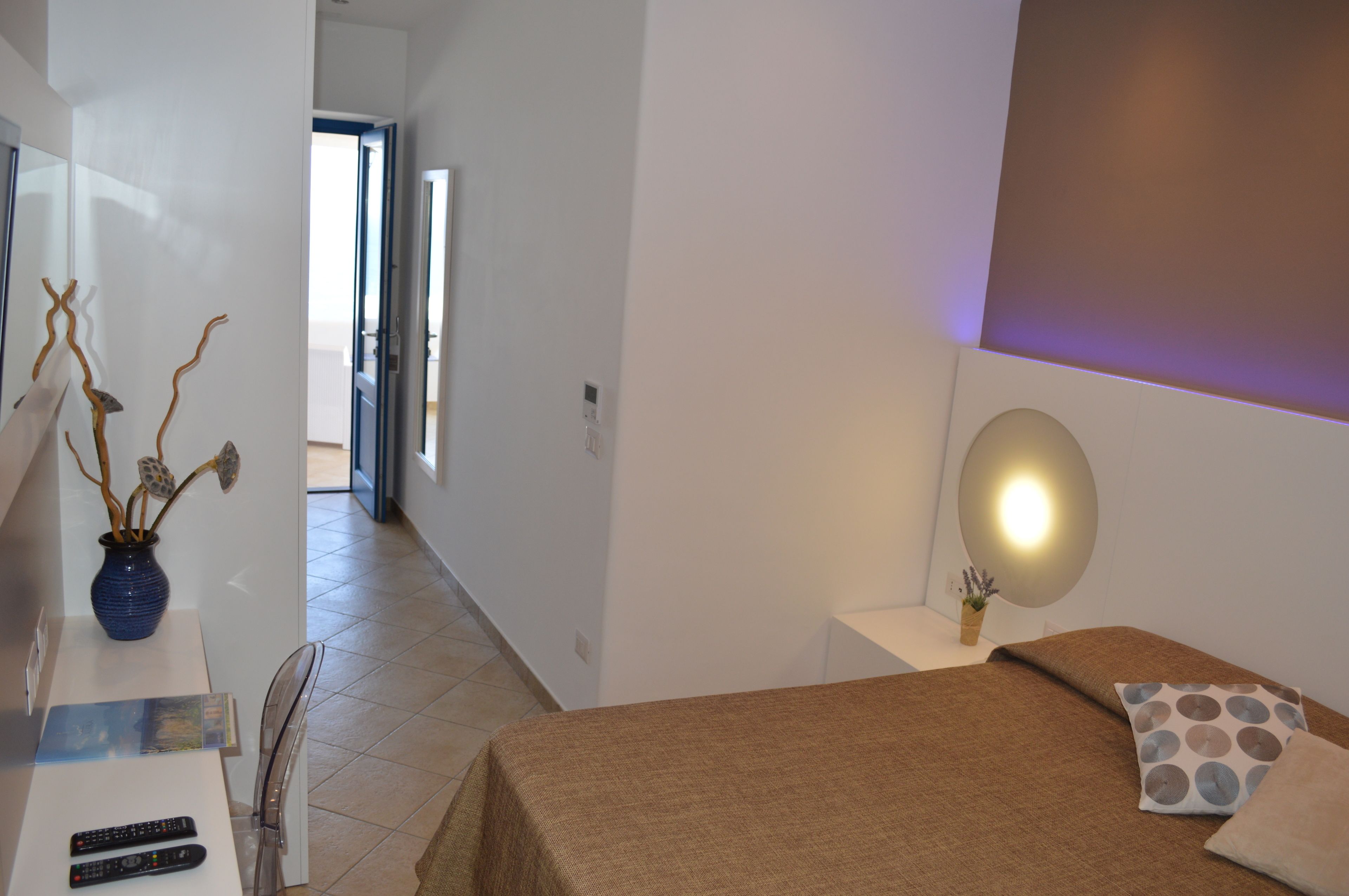 A hotel room with a bed, a bedside table with a lamp, a vase with branches, and a view of the hallway.