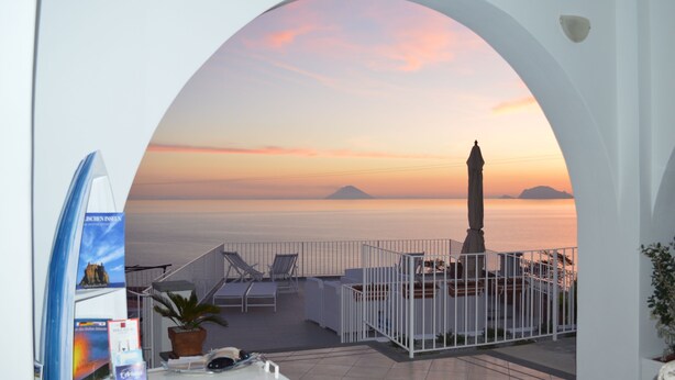 A terrace with a view of the sea at sunset, featuring a statue, lounge chairs, and a white railing. - A Alera Room and Breakfast (Malfa)